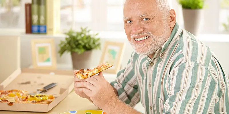 älterer Mann isst Pizza mit falschem Lachen – Sinnbild für kognitive Dissonanz in der Ernährung