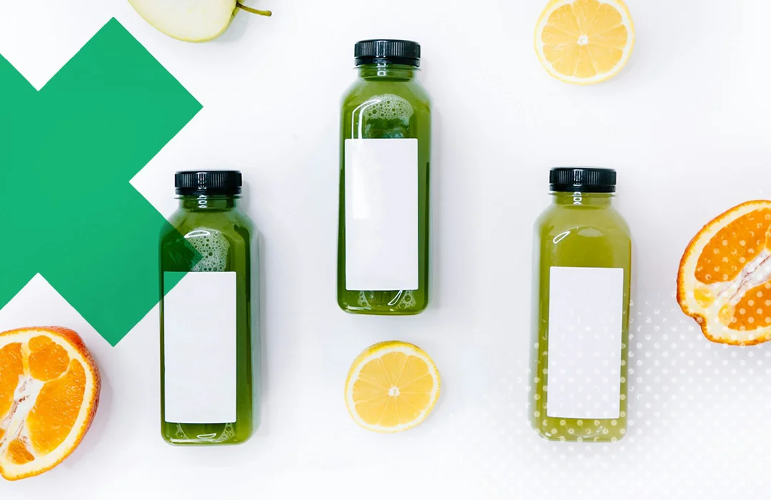 3 Smoothie Flaschen mit grünem Saft und weißem, leeren Etikett, umgeben von Orangen und einem aufeschnittenen Apfel